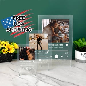 May include: Three clear acrylic photo frames with a music player design. Each frame has a different photo of a couple. The frames are on a stand and have a heart icon, play, pause, and rewind buttons. The text on the frames reads "Your Song Title Here", "Your Artist Name Here", "Song Title Here", "Artist Name Here", "Happy Anniversary Bift, I love You".