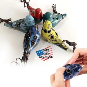 May include: A collection of ceramic ocarinas in the shape of birds. The ocarinas are in various colors, including blue, red, green, and yellow. Each has a brown cord attached. The image also includes the text "FREE USA SHIPPING".