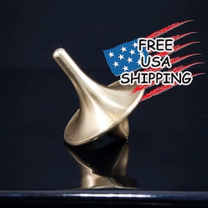 May include: A gold-colored metal spinning top with a pointed tip. The top is on a black surface.