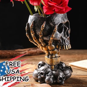 May include: A black and gold skull-shaped vase holding red roses. The vase is supported by a skeletal hand and a base of smaller skulls. A unique decorative piece for Halloween or gothic decor.