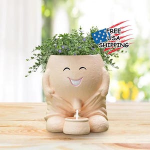 May include: A whimsical beige ceramic planter shaped like a smiling figure with a humorous design. The planter is filled with green foliage and small purple flowers. The planter has a small pot at the bottom.