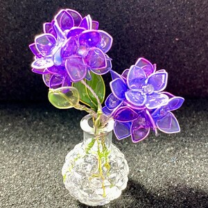 Könnte beinhalten: Eine kleine, klare Glasvase mit strukturierter Oberfläche enthält zwei lila Blüten. Die Blumen haben durchscheinende Blütenblätter und grüne Stiele. Die Vase steht auf einer dunklen, strukturierten Oberfläche.