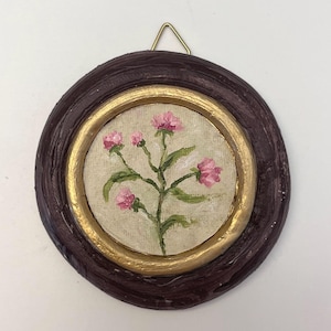 May include: A circular wall hanging featuring a small oil painting of a pink flower stem with green leaves. The artwork is encased in a textured dark brown frame with a gold inner rim and includes a small gold metal triangular hanging loop.