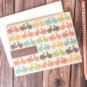 May include: A cream-colored thank you card with a repeating pattern of colorful bicycles. The word "thanks" is in a brown rectangle. A white envelope and a pink pencil are also in the image.