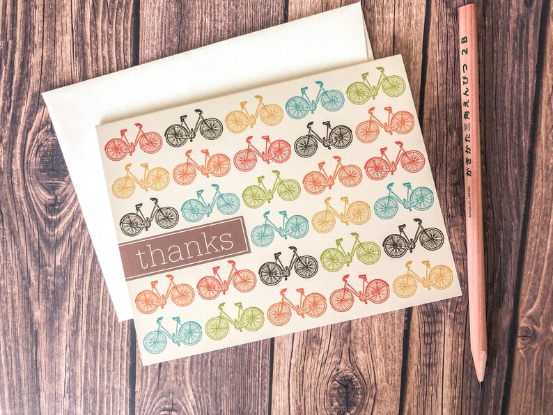 May include: A cream-colored thank you card with a repeating pattern of colorful bicycles. The word "thanks" is in a brown rectangle. A white envelope and a pink pencil are also in the image.