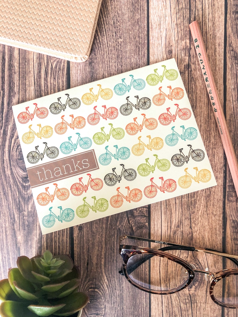 May include: A greeting card with a bicycle pattern in various colors, including red, green, and yellow. The card has the word "thanks" in a brown rectangle. A pencil, glasses, and a succulent are also visible.