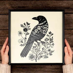 Op de afbeelding: Zwart-wit ingelijste kunstdruk met een gedetailleerde illustratie van een kraai die op een bloeiende tak zit. Het lichaam van de kraai is versierd met bloemmotieven en het kunstwerk is geplaatst tegen een crèmekleurige achtergrond.