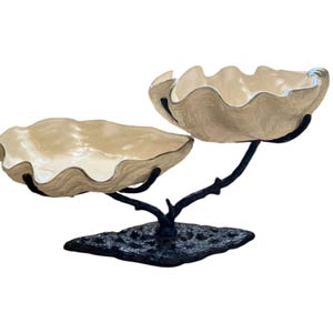 May include: A two-tiered decorative serving dish. The dish features two clam shell-shaped bowls in a cream color with a silver trim. The bowls are supported by a black metal branch-like structure and a textured base.