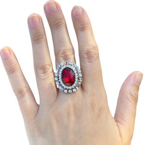 May include: A large, ornate ring featuring a vibrant red oval-cut gemstone at its center. The gemstone is surrounded by a halo of smaller, clear, round and baguette-cut stones, set in a silver-toned metal band. The ring is displayed on a hand.