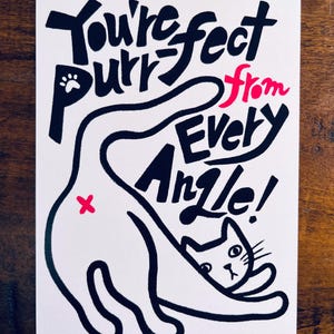 May include: A white card with a black and pink cat illustration. The card features the text "You're purrfect from every angle!" in a playful font. The cat is drawn in a simple, cartoon style.