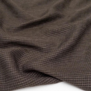 Può includere: Primo piano di un tessuto marrone con un sottile motivo a griglia. Il tessuto ha un aspetto strutturato con sottili linee bianche che creano un design a scacchi. Il materiale è drappeggiato, mostrando pieghe e ombre.