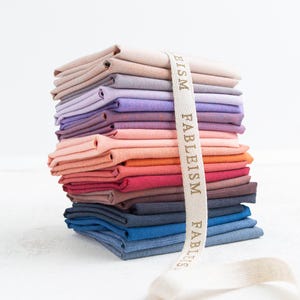 Pode incluir: Uma pilha de quadrados de tecido dobrados em várias cores, incluindo bege, lavanda, roxo, rosa, laranja, vermelho, azul marinho e azul. Uma fita creme com a palavra "FABLEISM" impressa envolve a pilha.