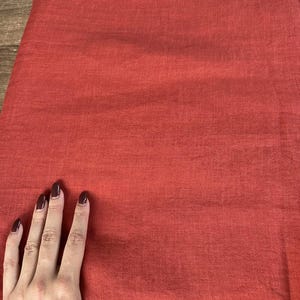 May include: A close-up of a folded, solid-colored fabric in a reddish-orange hue. The fabric has a textured appearance, with visible woven threads. The image shows a hand touching the fabric.