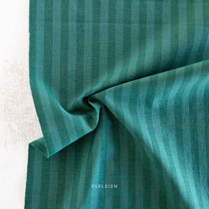 Può includere: Primo piano di un tessuto piegato con strisce verticali in varie tonalità di verde. Il tessuto ha un aspetto strutturato ed è parzialmente spiegazzato, rivelando l'intreccio. La parola "FABLEISM" è stampata in bianco in basso a destra.
