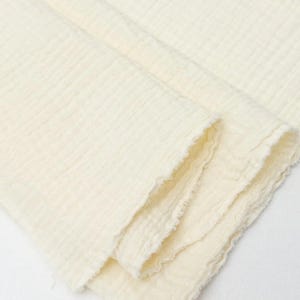 May include: A folded piece of off-white double gauze fabric featuring a crinkled, textured surface. The material displays a soft, layered appearance with raw, slightly frayed edges visible along the folds of the textile.