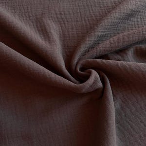 May include: A dark brown fabric with a distinct crinkled, double-gauze texture. The material is draped in soft, flowing folds, highlighting the uneven, puckered surface of the textile. The deep, earthy brown color is consistent across the entire piece.