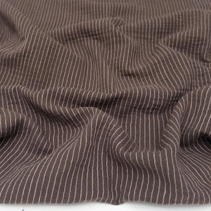 Op de afbeelding: Een donkerbruine stof met een patroon van dunne, verticale witte strepen. Het materiaal heeft een gekreukt, getextureerd oppervlak met zachte plooien en draperingen, wat een gelaagd uiterlijk creëert over het gehele textiel.
