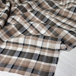May include: Close-up of a fabric with a plaid pattern in shades of brown, beige, black, and white. The fabric is folded and draped, showing the texture and weave of the material. The edges are visible, showing the fabric's construction.