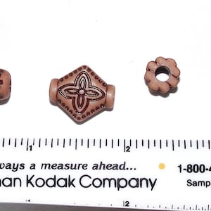 May include: Four brown plastic beads with different designs. The beads are about 1 inch long and have a variety of patterns, including a floral design, a diamond shape with a four-leaf clover, a circle with a hole in the center, and a bead with vertical lines.