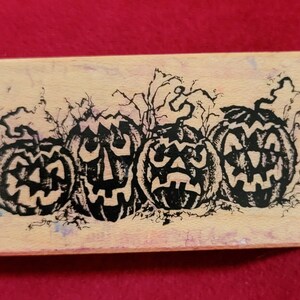 May include: A wooden stamp featuring a design of four black jack-o'-lantern faces with spooky expressions. The jack-o'-lanterns are surrounded by black vines and leaves.