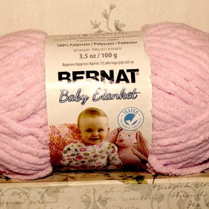 May include: A skein of soft, light pink Bernat Baby Blanket yarn with a paper label featuring the brand name, a photo of a baby, and text stating 100% polyester, 3.5 oz, and 72 yards. The yarn has a thick, chenille-like texture and is wrapped.