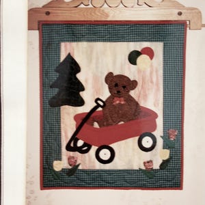May include: A quilted wall hanging featuring a brown teddy bear riding in a red wagon. The scene includes a black Christmas tree, balloons, and flowers. The quilt is framed with a green and red border and a wooden hanger.
