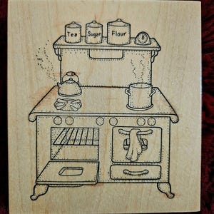 May include: A wooden stamp featuring a detailed illustration of a vintage kitchen stove. The stove has an open oven door, a pot, a kettle, and a shelf with jars labeled "Tea", "Sugar", and "Flour". A small clock is also on the shelf.