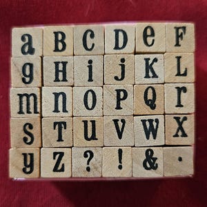 May include: A set of thirty square wooden stamps arranged in a grid, each featuring a black printed character. The collection includes the lowercase and uppercase alphabet letters from a to Z, along with a question mark, exclamation point, ampersand, and period.