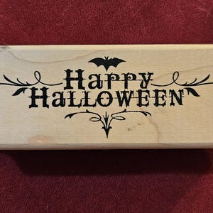 May include: A rectangular wooden rubber stamp featuring the black text "Happy Halloween" in a decorative font. A small black bat silhouette is positioned above the text, and ornate black flourishes extend from both sides and below the lettering.