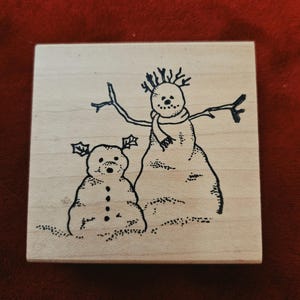 May include: A square wooden rubber stamp featuring a black ink illustration of two snowmen. The taller snowman wears a scarf and has twig arms, while the smaller snowman has holly leaves on its head. Both figures are depicted with dotted textures.