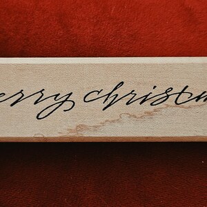 May include: A rectangular light-coloured wooden block featuring the words Merry Christmas written in a black cursive script font. The wood grain is visible across the surface of the block, which has a smooth, flat finish and straight edges.