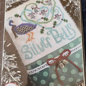 May include: A cross-stitch pattern titled Silver Bells featuring a peacock with purple and brown plumage, light blue swirls, and green holly leaves. The lower section has green polka-dot fabric, white lace trim, and a red bow with two small silver bells.
