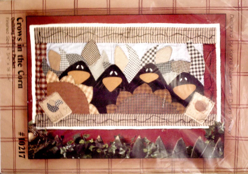 K&ouml;nnte beinhalten: Ein Quilt-Musterpaket mit dem Titel Crows in the Corn, #10217, mit einer fertigen Gr&ouml;&szlig;e von 86,36 cm mal 45,72 cm. Das Design zeigt vier schwarze Kr&auml;henfiguren mit orangefarbenen Schn&auml;beln, umgeben von karierten und gemusterten Stoffst&uuml;cken.