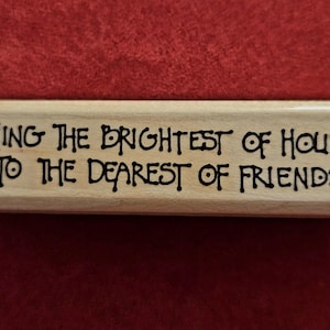 May include: A rectangular light-coloured wooden rubber stamp featuring the black engraved text: WISHING THE BRIGHTEST OF HOLIDAYS TO THE DEAREST OF FRIENDS, flanked by two small heart symbols with dots underneath on the left and right sides.