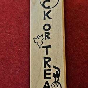 May include: A rectangular light-coloured wooden bookmark featuring the vertically engraved black text TRICK OR TREAT. The design includes a circular moon with a bat, a small ghost, and a black cat sitting atop a carved jack-o'-lantern pumpkin.