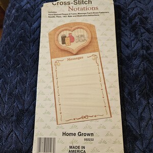 May include: A Cross-Stitch Notations craft kit package titled Home Grown. The packaging features a heart-shaped frame design, a message pad area, and lists included items such as a hand-stained plaque, frame, message pad, brass fasteners, needle, floss, and 14ct Aida cloth.