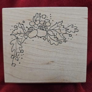 Puede incluir: Un sello de madera con un dibujo lineal negro de bellotas, hojas y bayas. El diseño está en la esquina superior izquierda del sello. El sello es de madera de color claro.