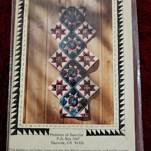 Puede incluir: Un paquete con un patrón de edredón para un camino de mesa. El patrón incluye instrucciones para la construcción de bloques. El diseño del camino de mesa presenta bloques en forma de diamante con patrones de estrellas en rojo, azul y blanco. El paquete está etiquetado como "Susie's Rising Star".