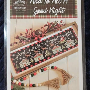 May include: A cross stitch pattern booklet titled And To All A Good Night by Stitching With The Housewives. The cover features a framed embroidery of a winter scene with houses, reindeer, and text, accented by red berries and wooden beads.