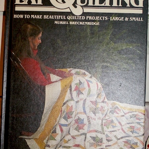 May include: Book cover titled "LAP QUILTING" with text "HOW TO MAKE BEAUTIFUL QUILTED PROJECTS - LARGE & SMALL" and author's name. The cover features a woman in a red top, seated, with a colorful quilt draped over her lap. The background is dark.