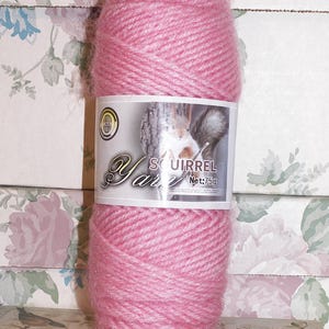 May include: A large spool of pink Squirrel Yarn, weighing 75g, is the focal point. The yarn is wrapped with a white label featuring the brand name and a squirrel image. The background is a floral pattern.