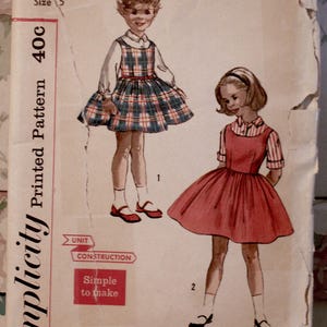 May include: Vintage Simplicity pattern #2209 for a child's dress, size 5. The illustration shows two children in dresses. One dress is plaid, the other is solid red. The pattern includes the text "Simple to make".