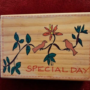 Könnte beinhalten: Ein rechteckiger Holzstempel mit einer handgemalten Illustration von zwei kleinen Vögeln auf einer Ranke mit einer großen orangefarbenen Blume. Der Text SPECIAL DAY ist in Rot aufgedruckt und von einem gestrichelten Rand umgeben.