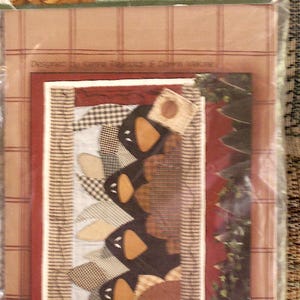 K&ouml;nnte beinhalten: Ein Quilt-Musterpaket mit dem Titel Crows in the Corn von Just Be Cuz Cousins in Design. Die Verpackung zeigt eine grafische Illustration von schwarzen Kr&auml;henformen mit orangefarbenen Schn&auml;beln, die vertikal vor einem karierten Hintergrund angeordnet sind.