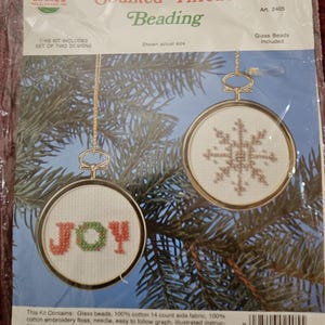May include: A set of two counted thread beading kits for Christmas ornaments. The kits include glass beads, 14 count aida fabric, embroidery floss, a needle, a graph, instructions, and gold metallic thread. The ornaments are a snowflake and the word "JOY".