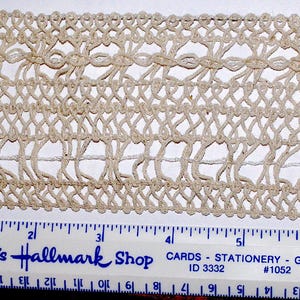 May include: Close-up of a beige lace trim with an open, woven design. The trim is measured with a ruler, showing a width of approximately 2 inches. The text "DeDe's Hallmark Shop" is visible.