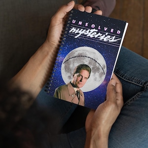 May include: A black spiral bound notebook with the words "UNSOLVED mysteries" in pink and white letters. The cover features a man in a brown coat standing in front of a large moon with a starry background. The text "Join me. Perhaps you may be able to help solve a mystery" is written in a circle around the moon.