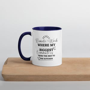 Peut inclure: Une tasse en céramique blanche avec un bord bleu. La tasse a un texte noir qui dit "Remote Work Where My Biggest Commute Is From The Bed To The Kitchen".