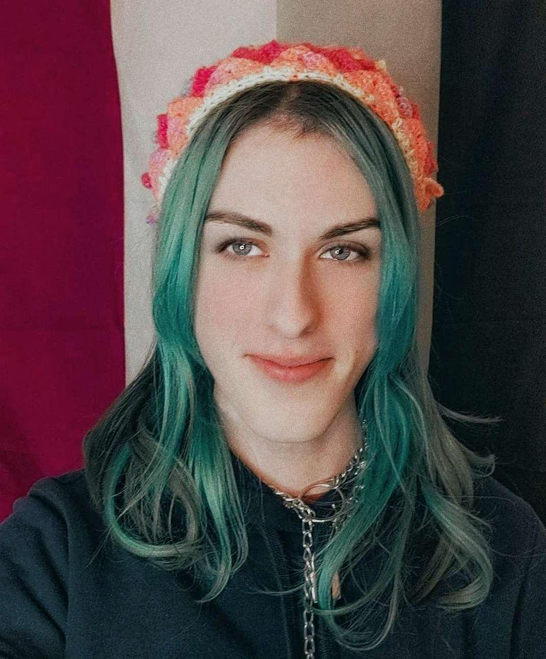 K&ouml;nnte beinhalten: Eine Person mit langen t&uuml;rkisfarbenen Haaren tr&auml;gt einen schwarzen Pullover und ein geh&auml;keltes Stirnband mit rosa, orangefarbenen und wei&szlig;en Akzenten.