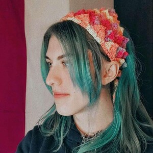 K&ouml;nnte beinhalten: Eine Person mit blaugr&uuml;nem Haar tr&auml;gt ein geh&auml;keltes Stirnband in den Farben Pink, Orange und Gelb. Das Stirnband ist am Hinterkopf zu einem Knoten gebunden. Die Person tr&auml;gt einen schwarzen Kapuzenpullover mit dem Wort "Jack" auf der Vorderseite.